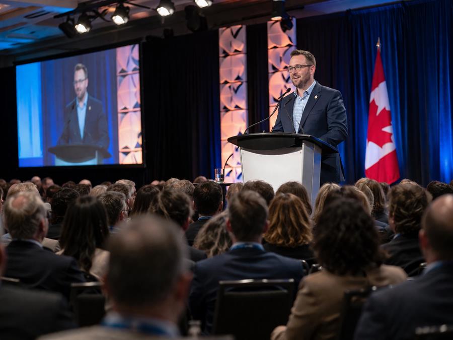 Mark McKenzie speaking at a podium to a full conference ballroom, with a Canadian flag on stage